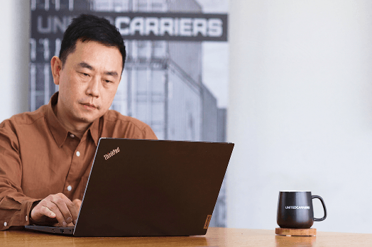 Man in brown shirt working on a ThinkPad laptop at a table with a black mug labeled Tidewell.